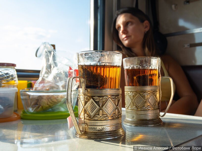 На столе в купе поезда стоят стаканы с чаем в металлических подстаканниках, на дальнем плане девушка смотрит в окно