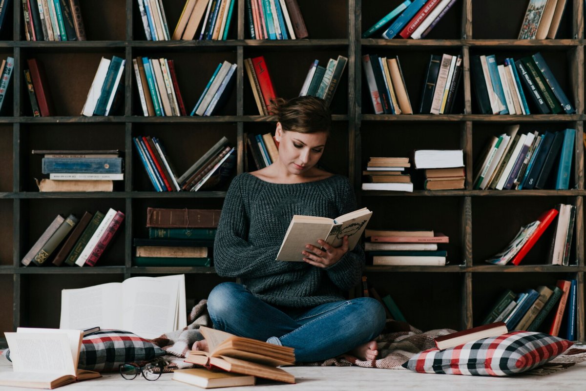 Девушка читает книгу, сидя рядом с книжным стеллажом